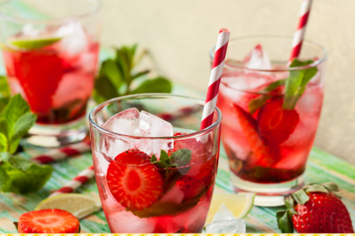 3 Gläser Erdbeer-Shrub mit Eiswürfeln, frischen Erdbeeren, Minze und Strohhalmen sind umgeben von Erdbeeren, Limetten und Minze. 