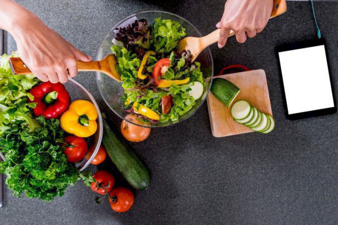 Eine Schale frischer Salat steht neben einer Schale mit den einzelnen Zutaten wie Blattsalat, gelbe und grüne Paprika, Petersilie, Paradeiser und Zwiebeln. Im fertigen Salat wird umgerührt und es liegt ein Tablett, das gerade aufgeladen wird daneben.