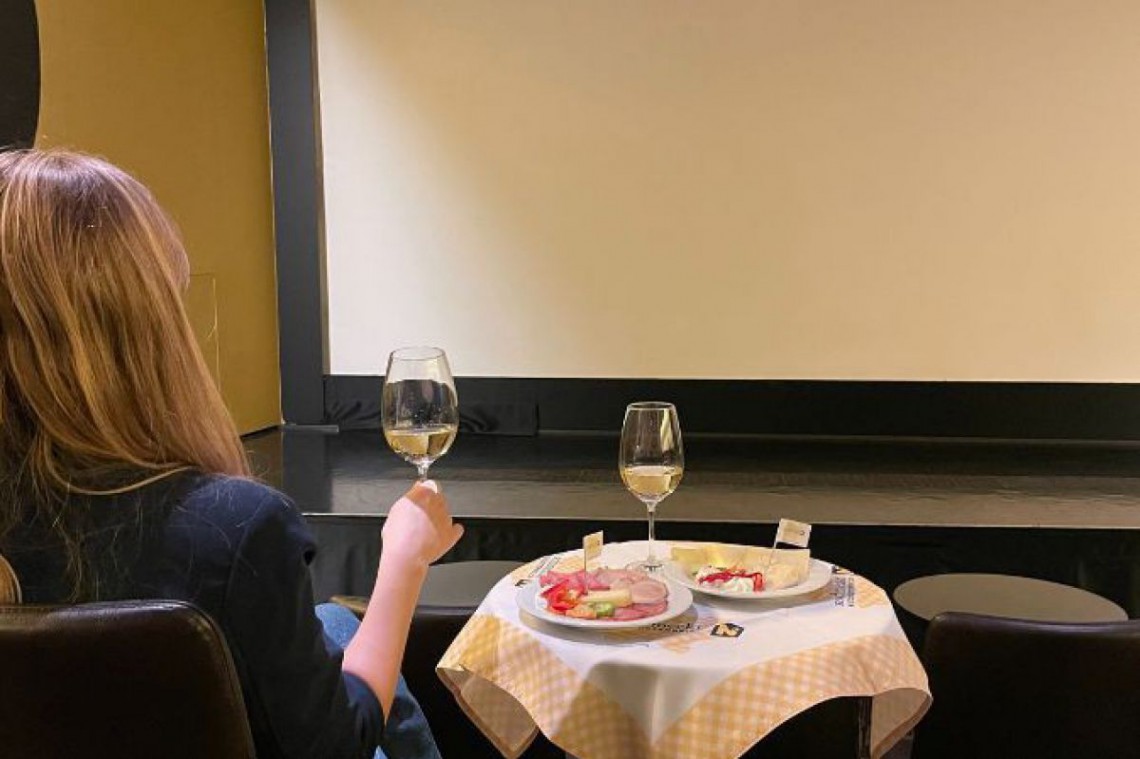 Dame sitzt mit einem Glas Wein und einem Teller voll mit regionalen Schmankerl vor der Kinoleinwand.