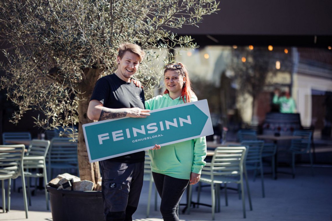 Besitzer von Feinsinn Genusslokal mit Schild in der Hand