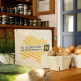 Bild anzeigen: Milchkasino Tisch mit Zwiebeln, Erdäpfeln, Lauch und Schnittlauch und der So-Schmeckt-Niederösterreich-Plakette 