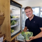 Bild anzeigen: Pepi Omas Speis, Herr Burger mir Karfiol in der Hand vor dem Kühlschrank im Verkaufsladen