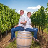 Bild anzeigen: Herr und Frau Greillinger sitzen auf Weinfass im Weingarten