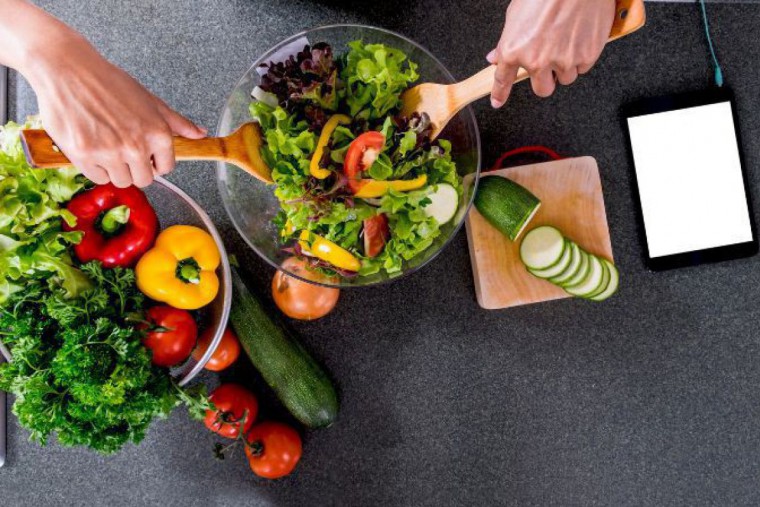 Eine Schale frischer Salat steht neben einer Schale mit den einzelnen Zutaten wie Blattsalat, gelbe und grüne Paprika, Petersilie, Paradeiser und Zwiebeln. Im fertigen Salat wird umgerührt und es liegt ein Tablett, das gerade aufgeladen wird daneben.