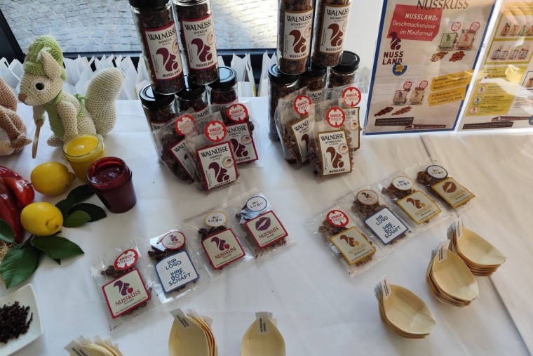 Stimmungsvolles Verkaufsdisplay für Nuss-Snacks von 'Nussland'. Auf einem weißen Tisch sind verschiedene Produkte ansprechend arrangiert: Gläser mit Walnüssen, kleine Snack-Packungen mit dem Eichhörnchen-Logo und informative Aufsteller. Handgestrickte Eic
